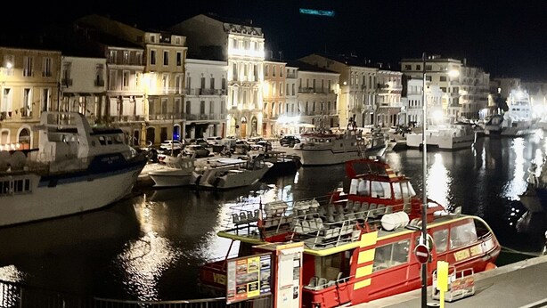 Un puerto nocturno con barcos amarrados, edificios con fachadas iluminadas y un bote rojo al frente.