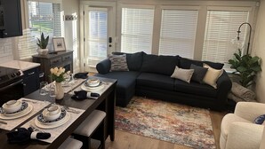 Een moderne woonkamer met een hoekbank, een eettafel voor vier personen en grote ramen met jaloezieën.