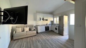 A modern kitchen with white cabinets, a built-in oven, microwave, and washing machine. There's a grey sofa and a large flat-screen TV mounted on the wall.