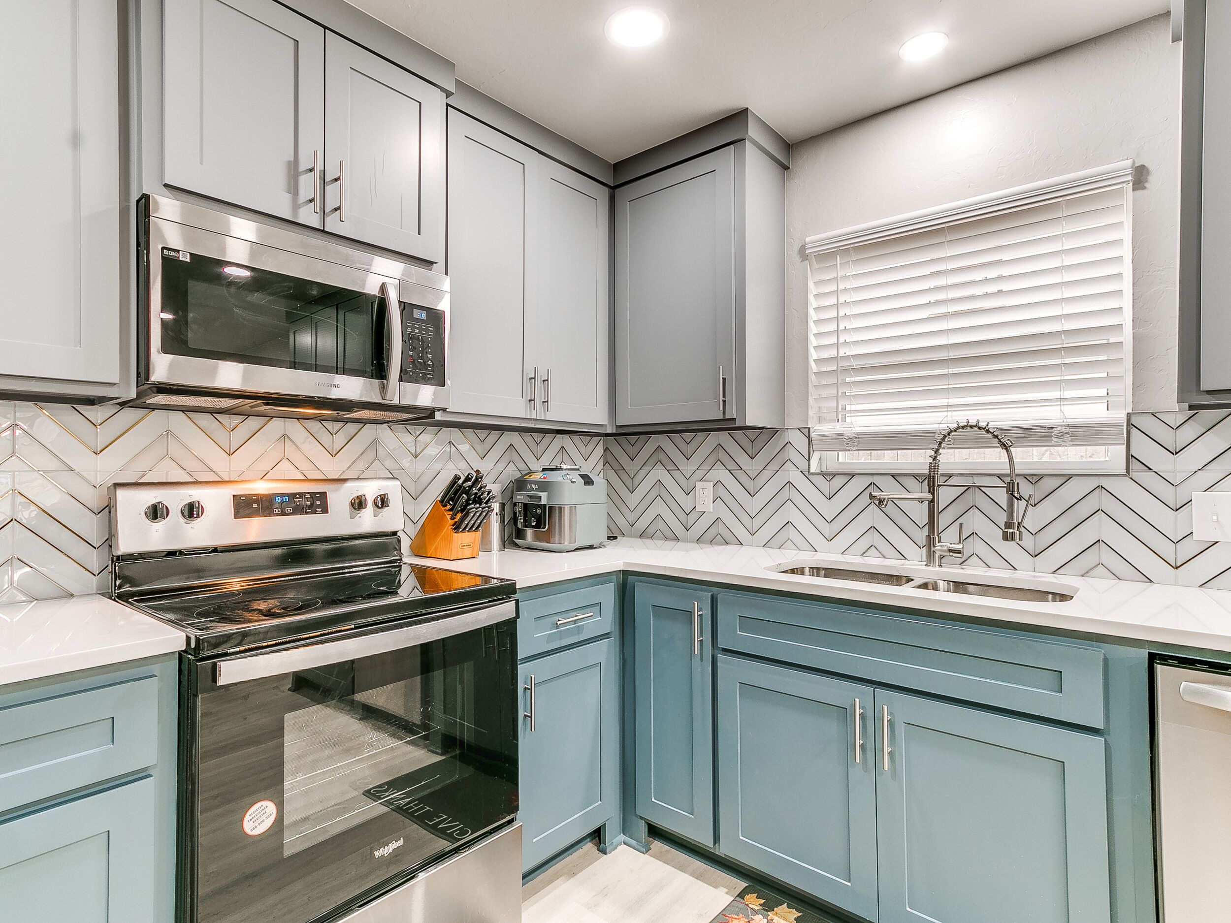 A modern kitchen with grey cabinets, a microwave, an oven, a sink, and a window with blinds.