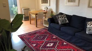 Una sala de estar con un sofá azul, un tapete rojo estampado, una mesa de comedor con sillas y un ventanal con vistas al exterior.