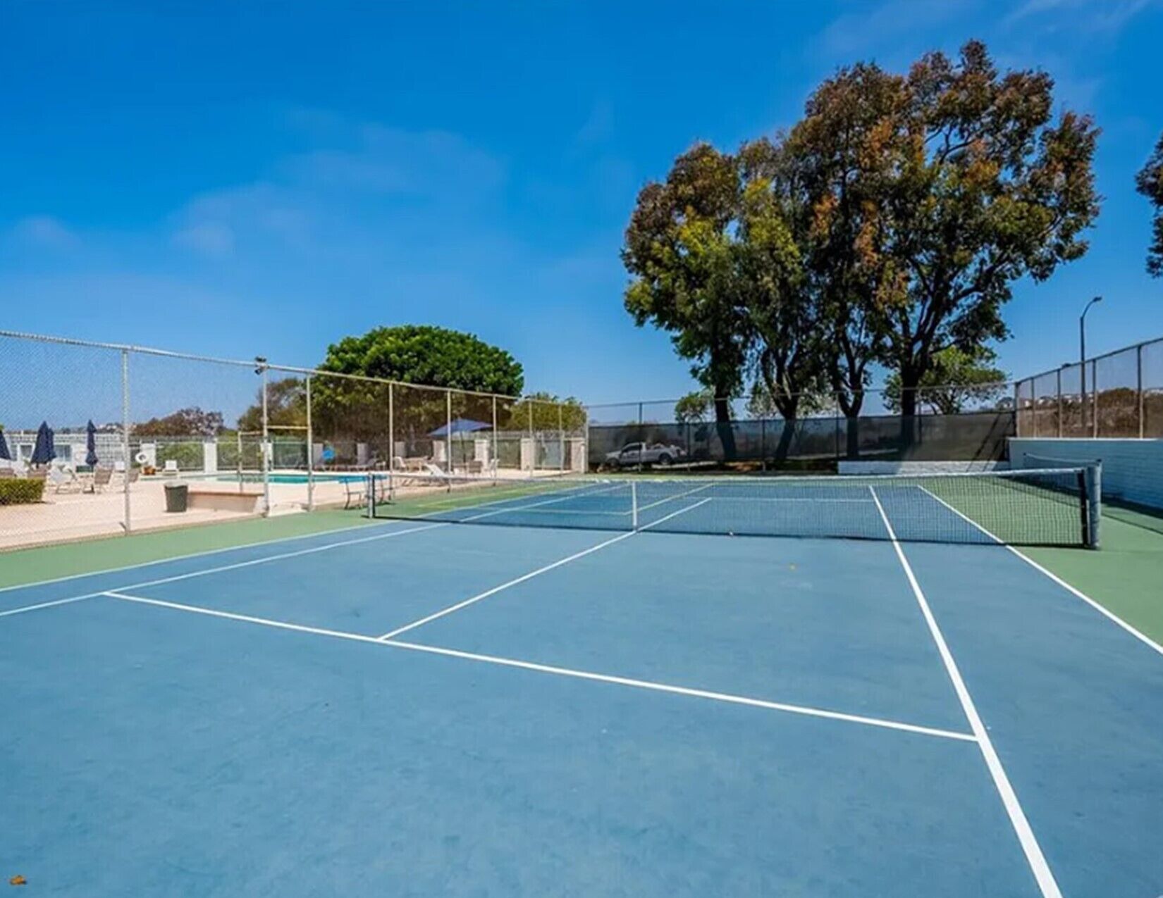Ein Tennisplatz mit Netz, umzäunt, sowie ein Poolbereich mit Liegestühlen und Sonnenschirmen.