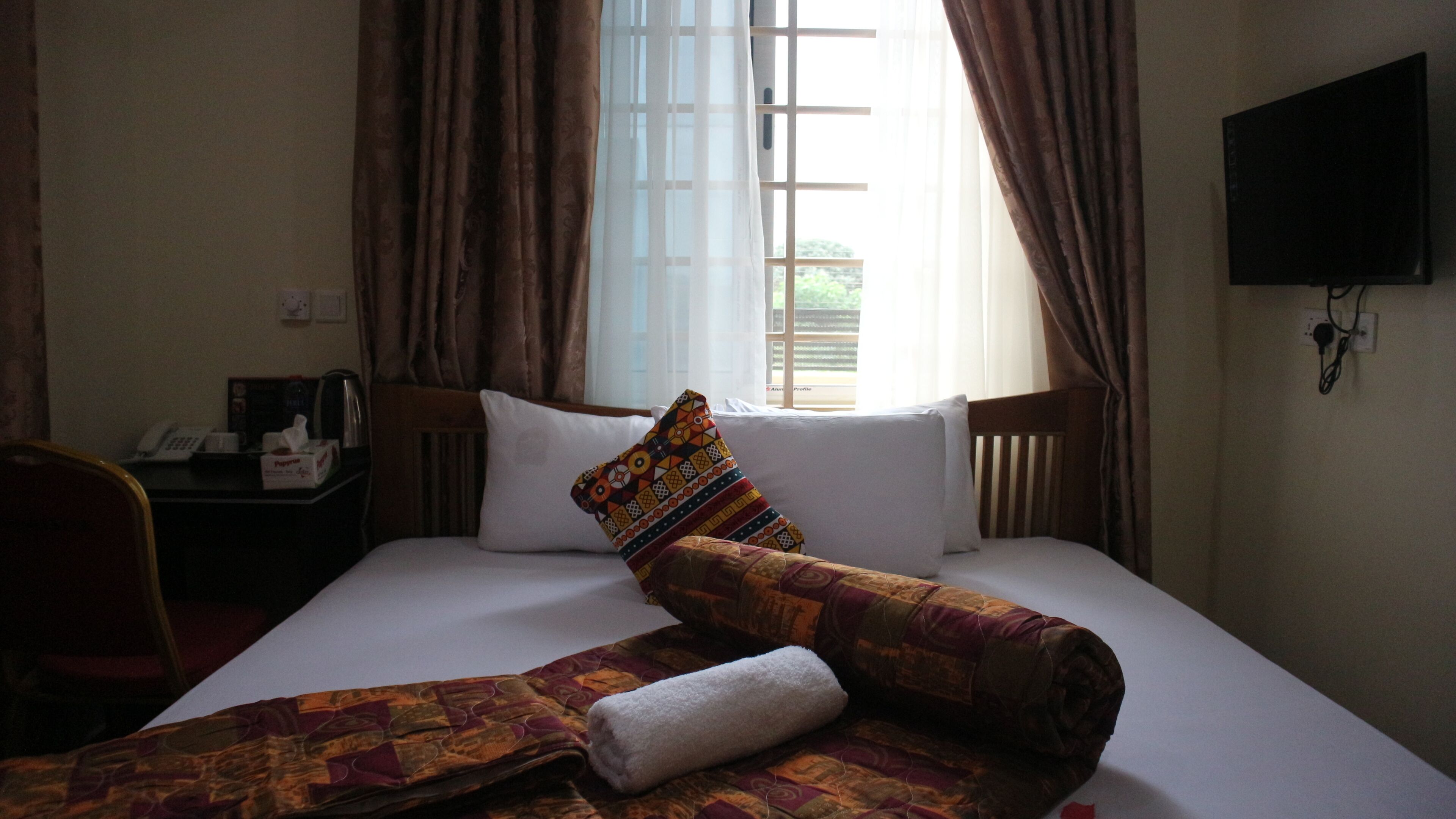 Una habitación de hotel con una cama, un televisor en la pared, un escritorio con teléfono y cafetera, y una ventana con cortinas.