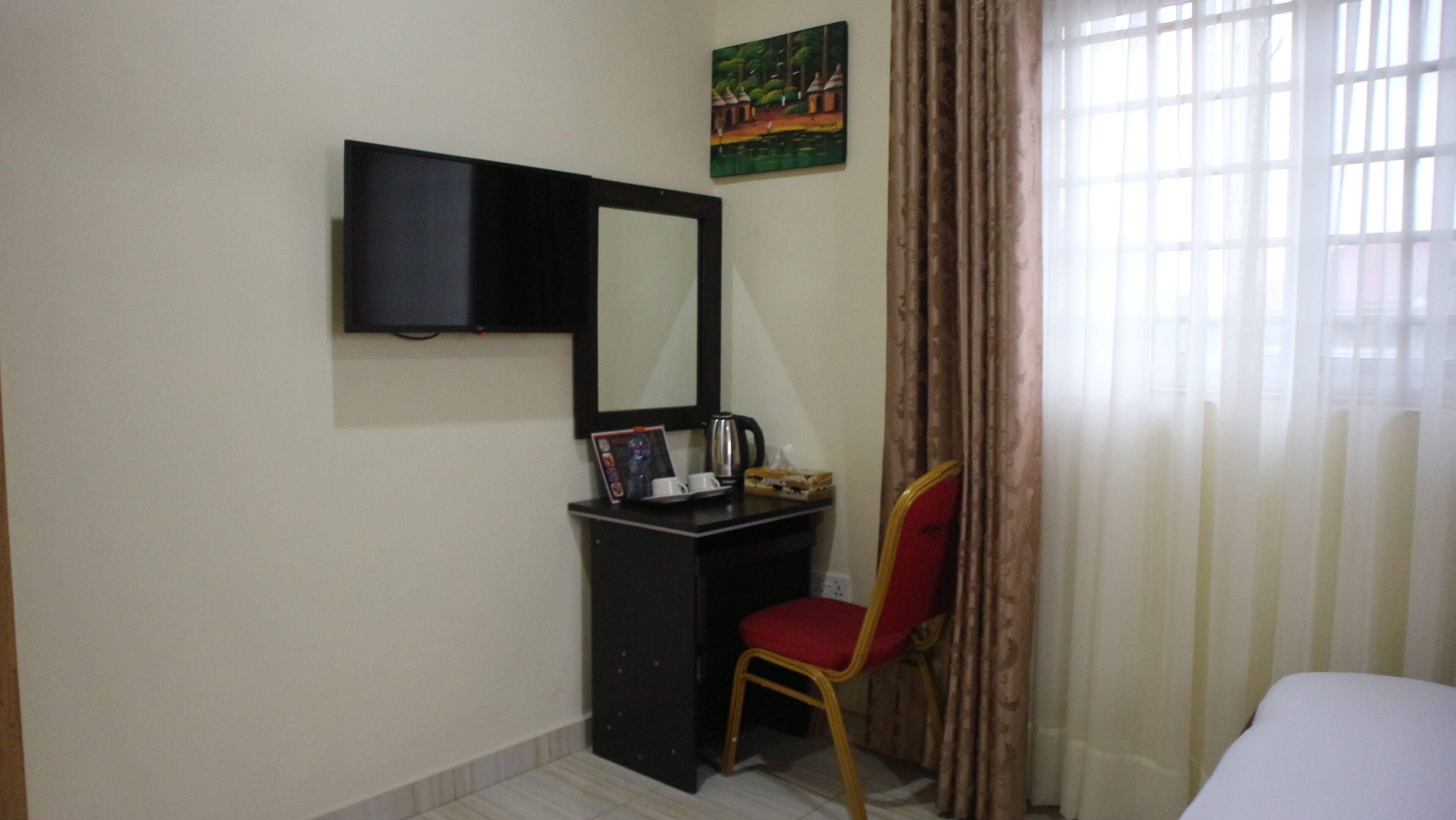 A hotel room with a bed, a desk with a chair, a television, a painting, and a window with curtains.
