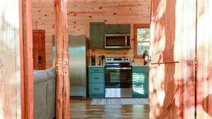 Ein gemütliches Blockhaushaus-Interieur mit Holzbalkendecke, einer Küchenzeile mit Mikrowelle, Herd und grünen Schränken sowie einem grauen Sofa.