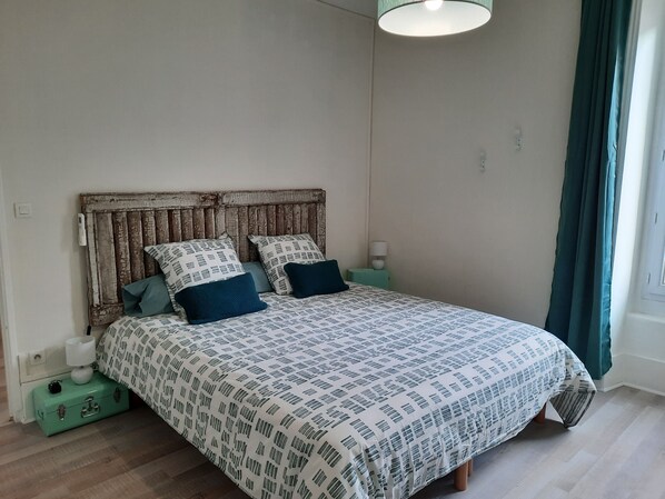 Une chambre à coucher avec un lit, une tête de lit, une table de chevet, une lampe, une valise et une fenêtre avec des rideaux.