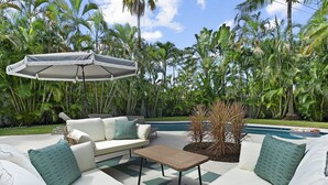 Ein Poolbereich mit weißer Gartenmöblierung, darunter eine Couch und ein kleiner Tisch, unter einem großen Sonnenschirm.