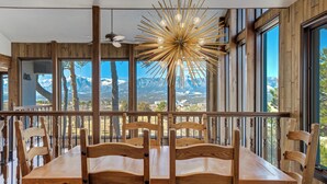 A dining area with a wooden table and chairs, large windows with a mountain view, and a unique ceiling light fixture.
