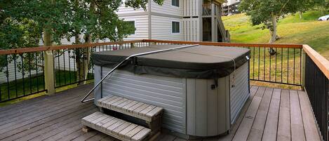 Ein Whirlpool auf einer Holzterrasse mit Metallgeländer, umgeben von Bäumen und einem Wohngebäude im Hintergrund.