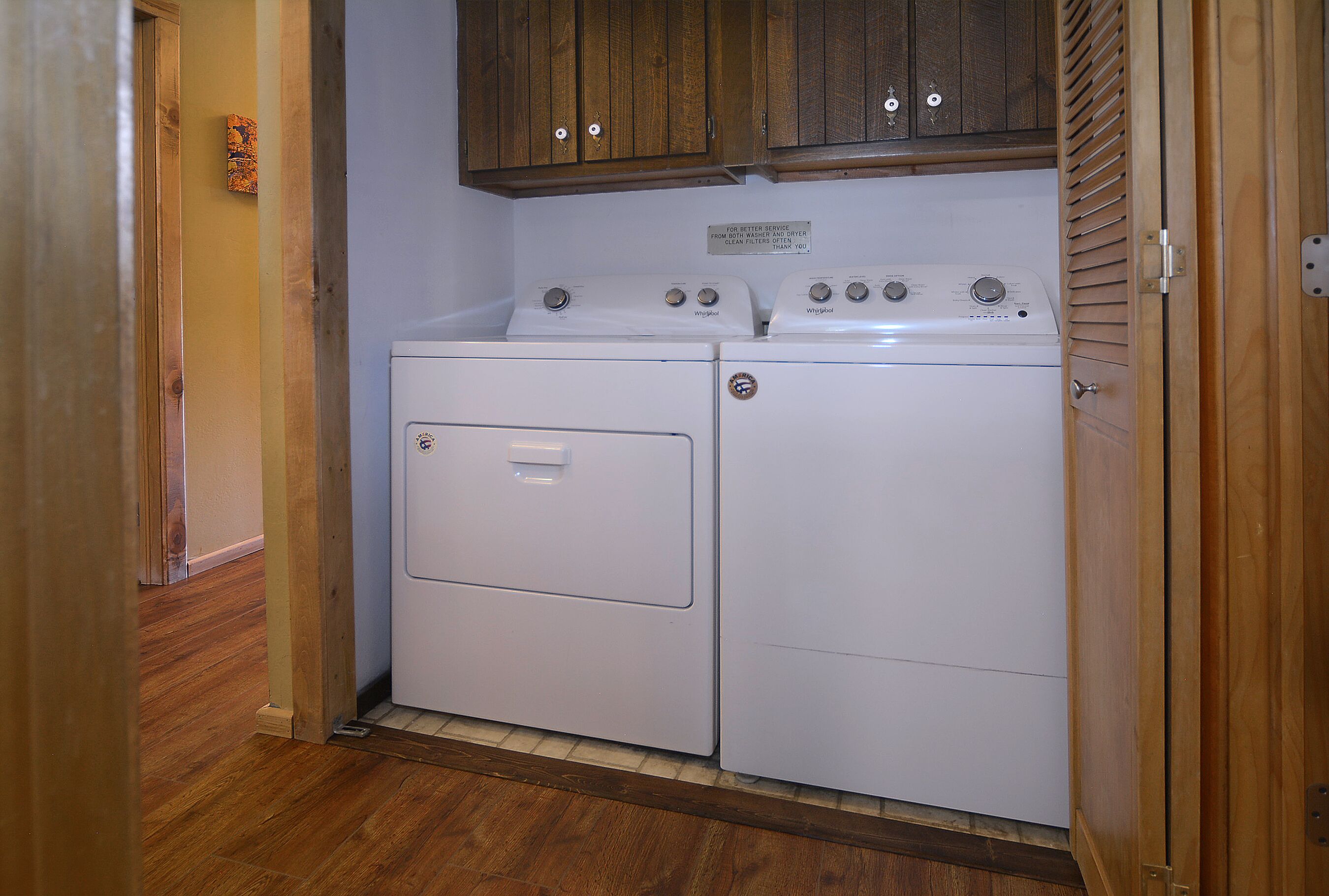 A laundry room with two white washers and dryers, wooden cabinets above, and a wooden floor.