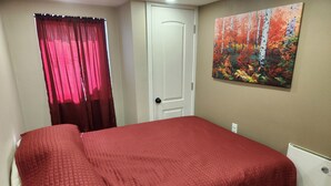 Un dormitorio con una colcha roja, una puerta blanca y un cuadro de un paisaje forestal en la pared.