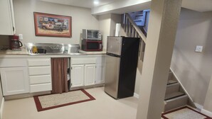 Cocina compacta con armarios blancos, refrigerador de acero inoxidable, microondas y tostadora. Se observa un cuadro en la pared y una escalera que sube a un nivel superior.