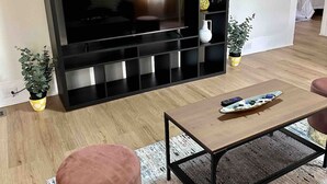 A modern living room with a large flat-screen TV mounted on a black entertainment unit, a coffee table, a pink ottoman, and two potted plants. - Beautiful contemporary 2BDR - Concord (Concord)