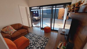 A modern living room with a brown sofa, orange ottoman, and a geometric rug. A wooden TV stand with a flat-screen TV and decorative items is visible. - Sabe Aquele Lugar Tranquilo e Lindo?! Assim é a Casa de Temporada Analu (Olímpia)