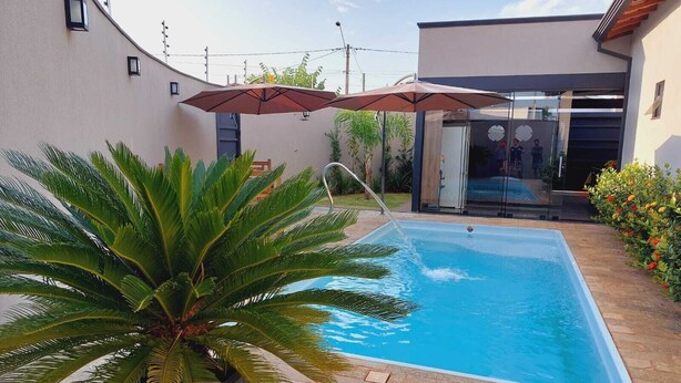 A swimming pool with a stone patio, surrounded by a modern house with glass doors and a large umbrella. - Sabe Aquele Lugar Tranquilo e Lindo?! Assim é a Casa de Temporada Analu (Olímpia)