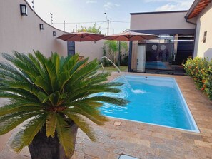 A swimming pool with a stone patio, surrounded by a modern house with glass doors and a large umbrella. - Sabe Aquele Lugar Tranquilo e Lindo?! Assim é a Casa de Temporada Analu (Olímpia)