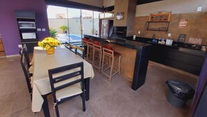 A modern kitchen with a bar area, a dining table with chairs, and a view of the outside. - Sabe Aquele Lugar Tranquilo e Lindo?! Assim é a Casa de Temporada Analu (Olímpia)