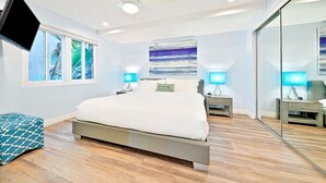 Ein modernes Schlafzimmer mit einem großen Bett, einem Deckenventilator, einem Flachbildfernseher und einem großen Fenster mit Blick auf Grünflächen.