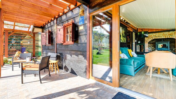 Una cabaña de madera con un patio amueblado con mobiliario de mimbre y un ventanal de vidrio que da a una sala de estar con chimenea.