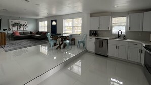 Un vaste espace de vie comprenant une table à manger, des chaises et un canapé. On peut apercevoir une cuisine avec des meubles blancs et un lave-vaisselle.