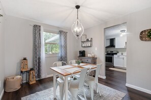 Un coin repas avec une table en bois, des chaises blanches, un lustre et une vue sur la cuisine.