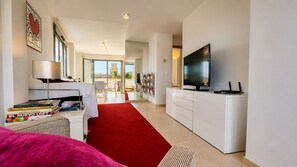A modern hotel room with a flat-screen TV, a red carpet, a dining area with a table and chairs, and a large window offering a view of the outside.