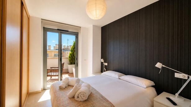 Una habitación de hotel moderna con una cama grande, un balcón con una silla y una planta, y una pared de acento oscura.