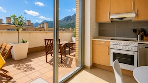 Una cocina moderna con armarios de madera, estufa y lavavajillas. Hay un balcón con mesa y sillas, y vistas a la montaña.