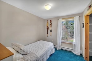 Quarto com uma cama de solteiro, janela com cortinas brancas e vista para árvores lá fora.