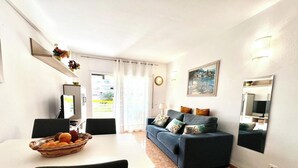 A living room with a sofa, a dining table with a fruit basket, a TV, and a painting on the wall. - Diamant Apartment, with parking and close to the beach. (salou)