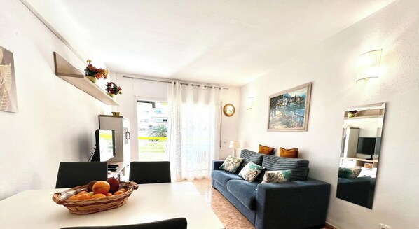 Uma sala de estar com um sofá, uma mesa de jantar com uma cesta de frutas, uma televisão e um quadro na parede.