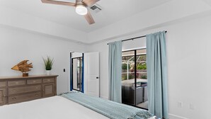 Ein Schlafzimmer mit Bett, Schminktisch, Deckenventilator, Fenster mit Vorhängen und Blick ins Freie.