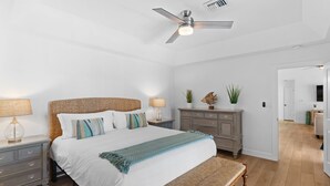 Ein Schlafzimmer mit Bett, Nachttischen, einer Bank, einem Deckenventilator, einer Kommode und einer Tür zu einem anderen Raum.