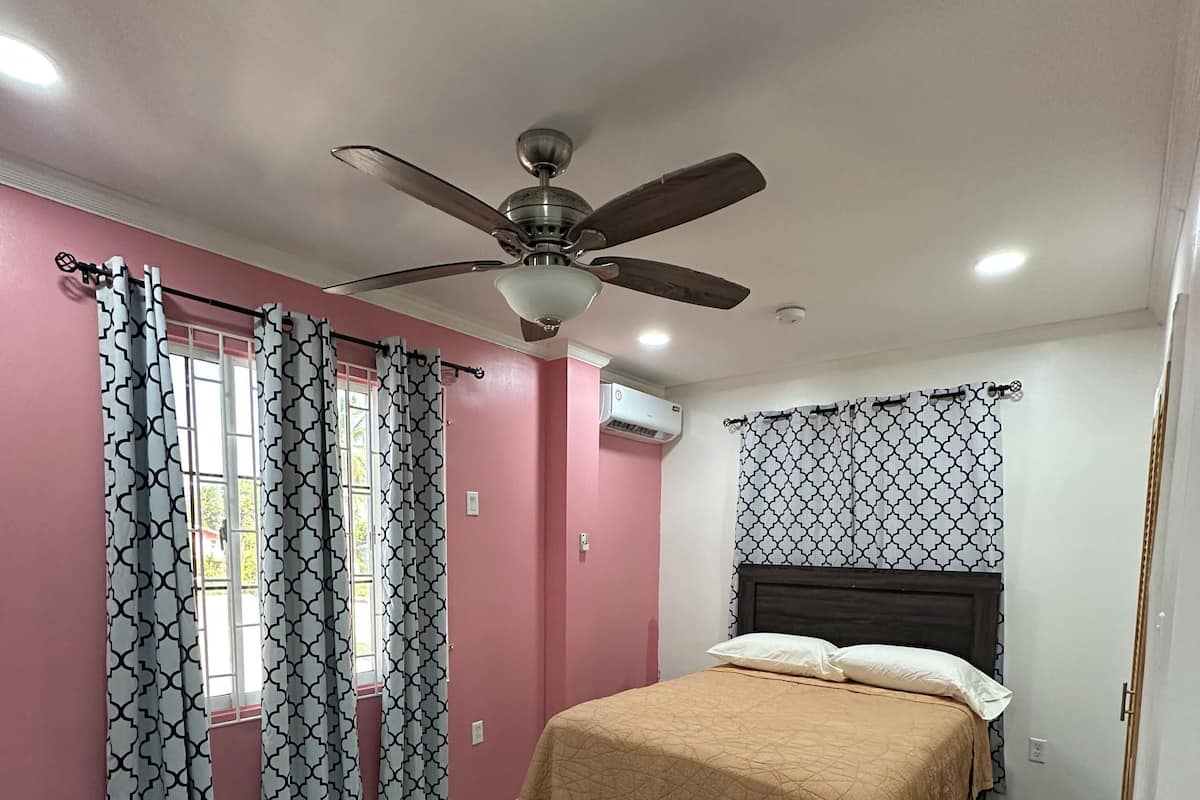 A bedroom with a bed, a ceiling fan, a window with patterned curtains, and an air conditioning unit.