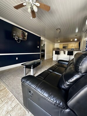 A modern living room with a black leather sofa, a glass coffee table, a ceiling fan, and a kitchen area in the background. - Your Next Level Int Executive Apartment (Georgetown)