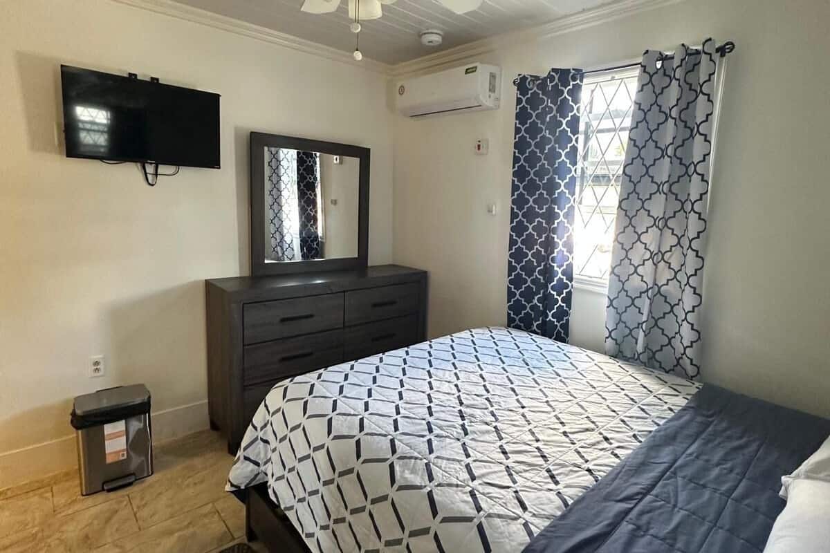 A bedroom with a bed, a ceiling fan, a TV, an air conditioning unit, and a window with patterned curtains.