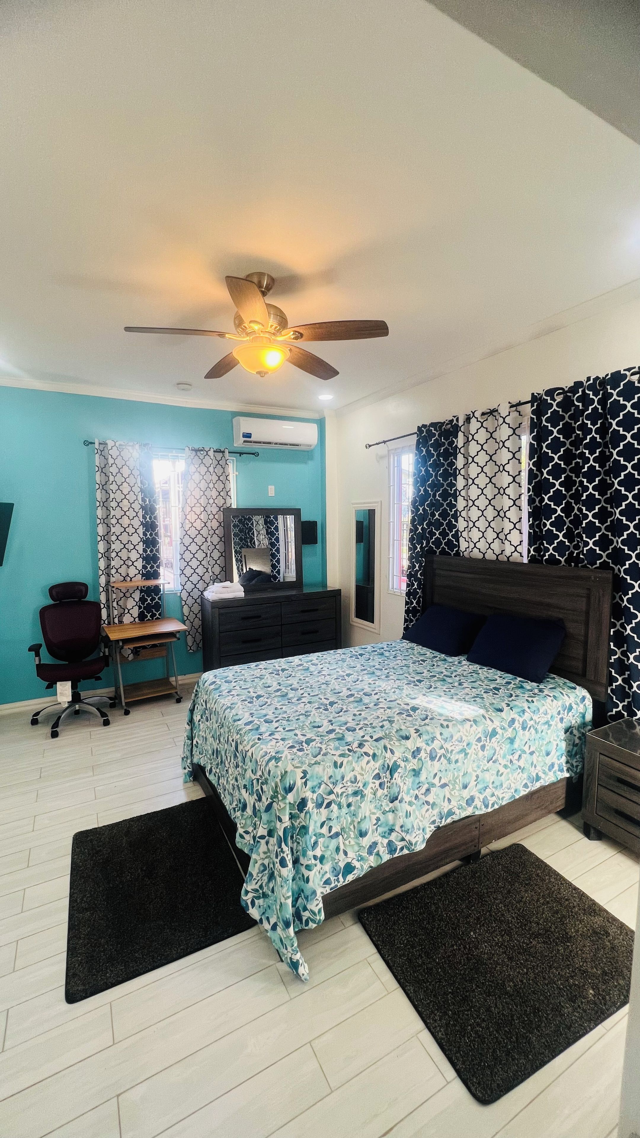 A bedroom with a bed, a desk, a chair, a ceiling fan, and patterned curtains.
