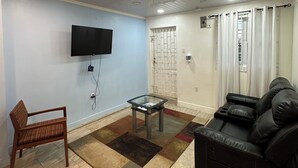 Un salón con una televisión de pantalla plana montada en la pared, una mesa de centro de vidrio, una silla de mimbre marrón, un sofá de cuero negro y una alfombra estampada.