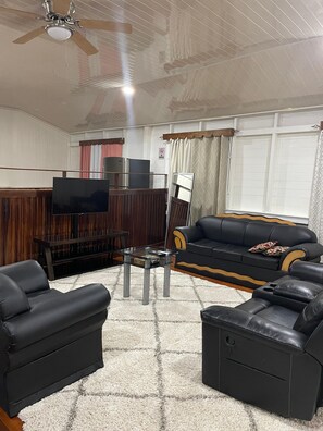 A living room with a flat-screen TV, a black leather sofa, a glass coffee table, and a ceiling fan. - Your Next Level Int Executive Apartment (Georgetown)