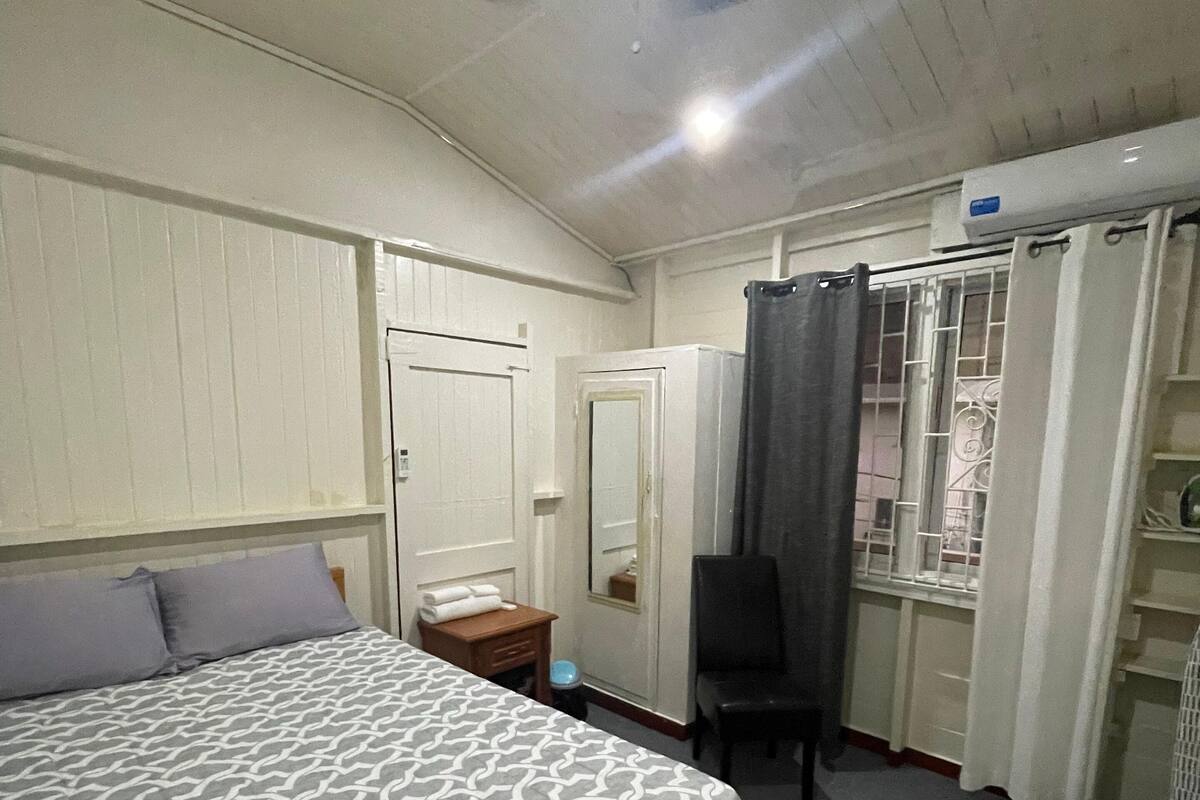 A hotel room with a bed, a chair, a desk, a mirror, a window with curtains, and a ceiling fan.