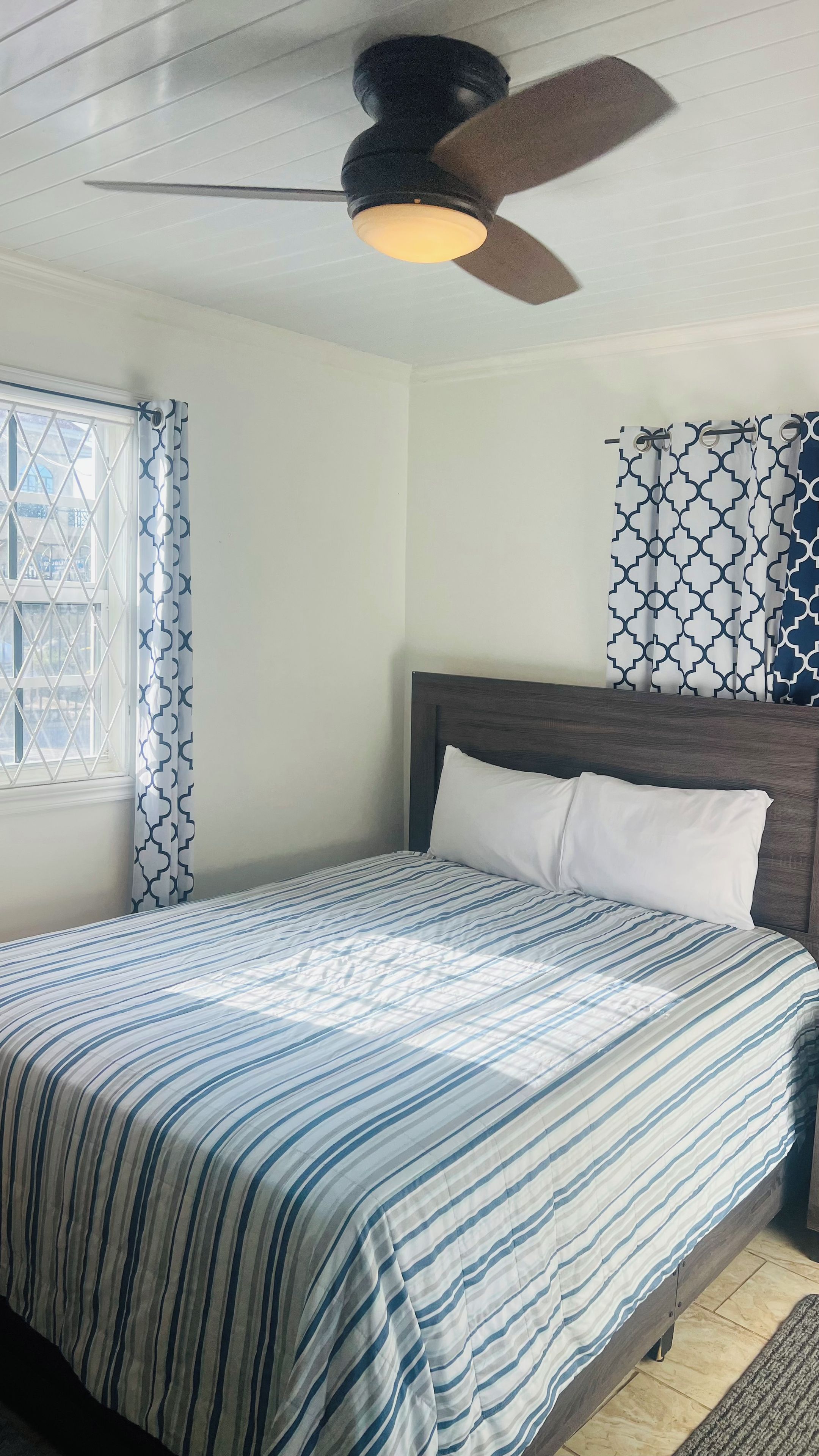 A bedroom with a bed, a ceiling fan, a window with patterned curtains, and a wall with a geometric-patterned curtain.
