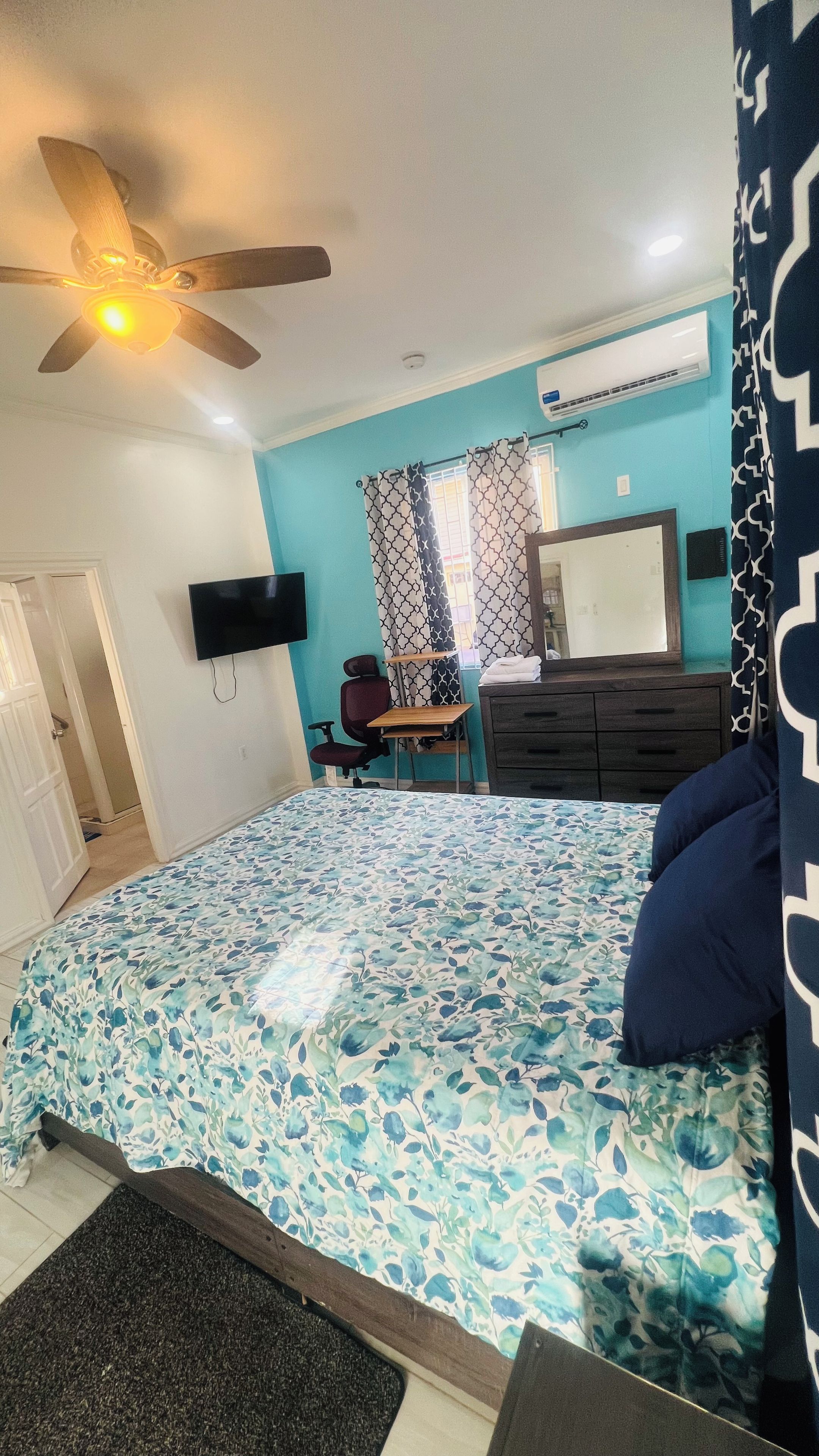 A bedroom with a bed, a desk, a chair, a TV, a ceiling fan, and an air conditioning unit.