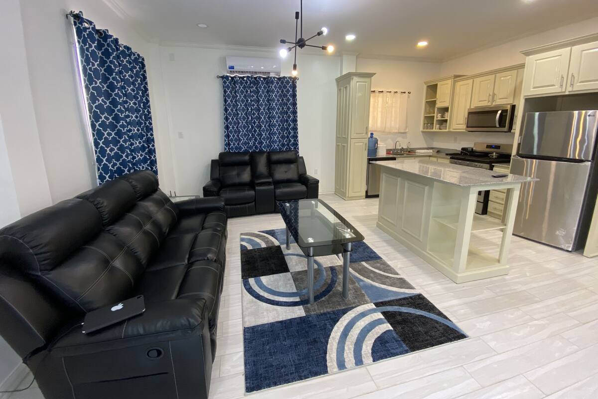 A modern living room with a black leather sofa and a glass coffee table, a kitchen with white cabinets and stainless steel appliances, and a patterned blue and white rug.