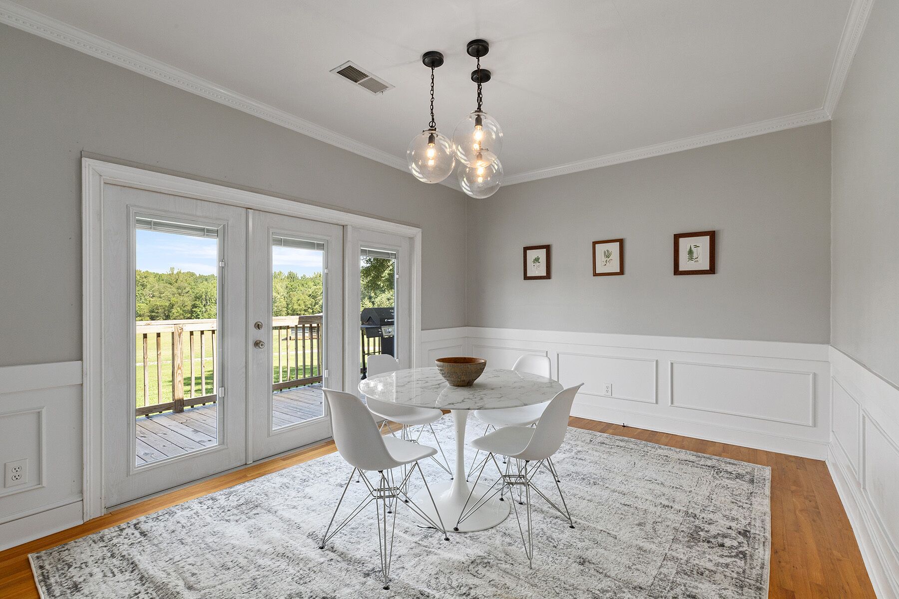 Un espace repas avec une table et des chaises blanches, un lustre et une vue sur une terrasse extérieure, accessible par des portes vitrées.
