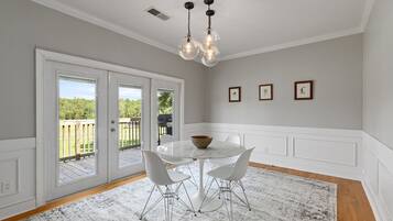 Un espace repas avec une table et des chaises blanches, un lustre et une vue sur une terrasse extérieure, accessible par des portes vitrées.