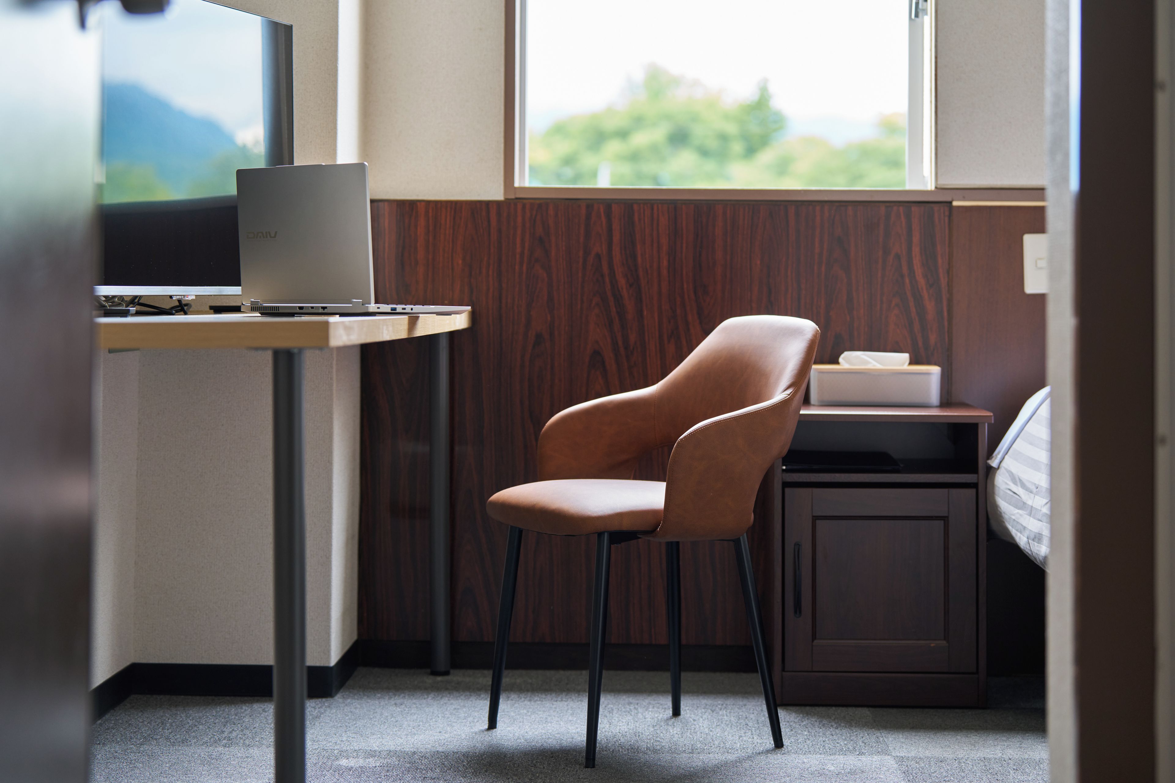 Een moderne kantoorruimte met een bureau, laptop, stoel en uitzicht op groen.