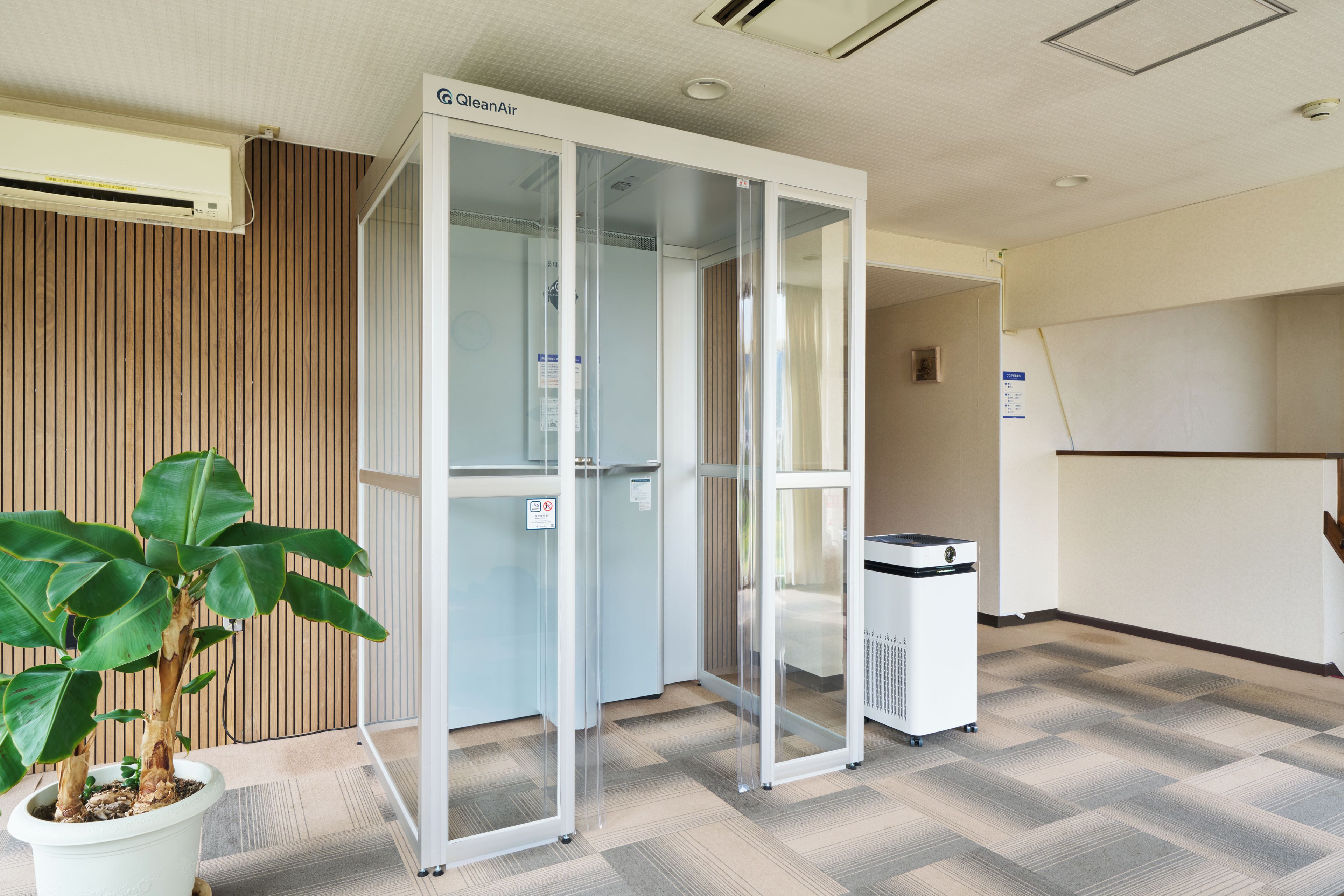 Un espacio de oficina moderno con un divisor de vidrio, una planta en maceta y un purificador de aire blanco.