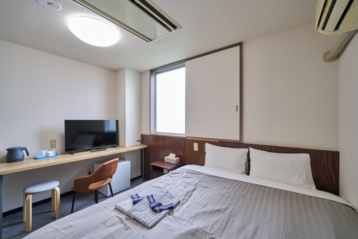 A hotel room with a bed, a desk with a television, a chair, and a window.