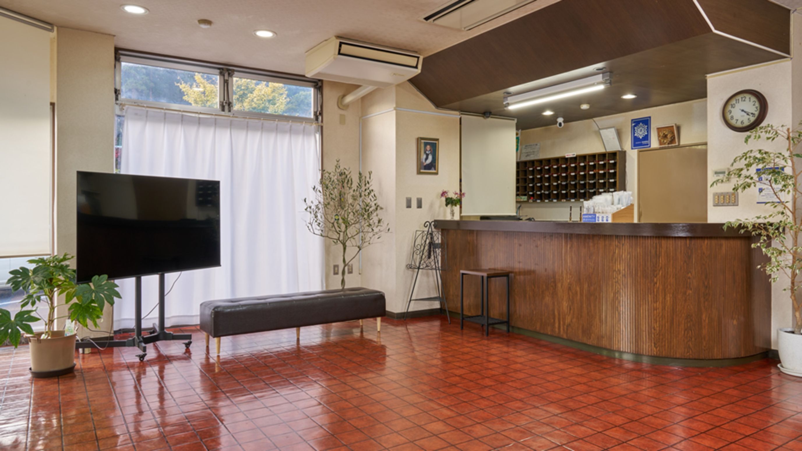 A reception area with a flat-screen TV, a wooden reception desk, a bench, a potted plant, and a clock on the wall.