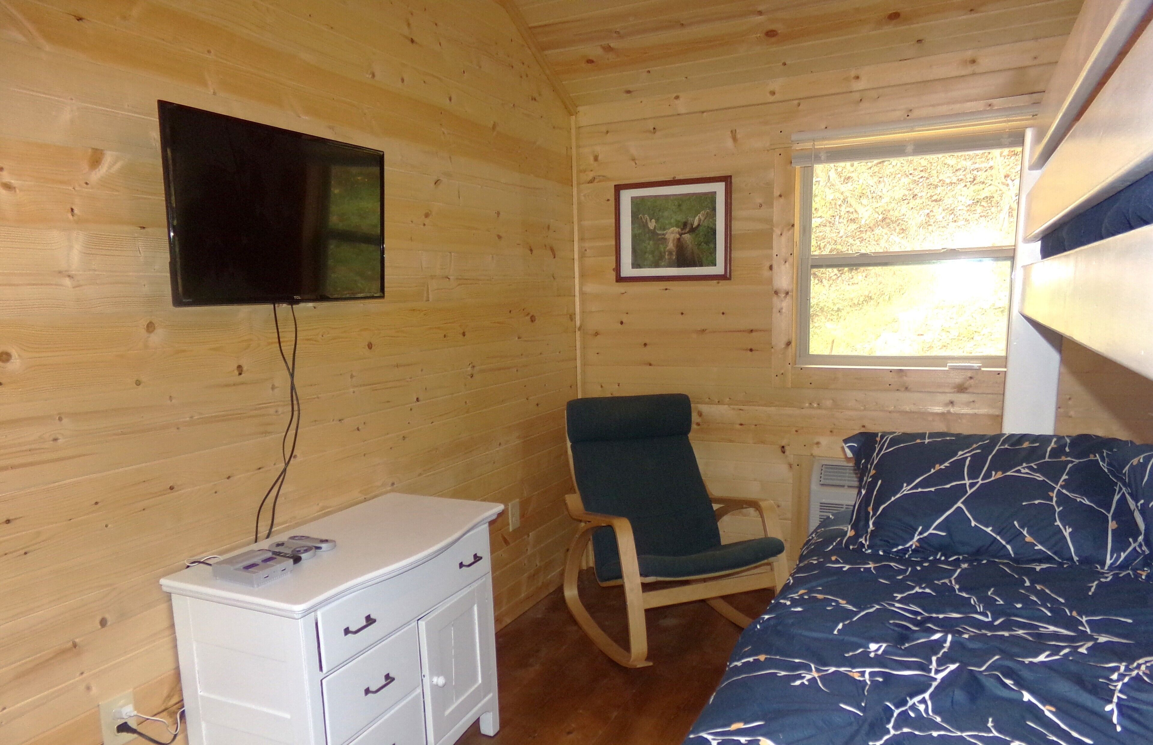 Een houten kamer met een bed, een schommelstoel, een dressoir, een aan de muur bevestigde televisie en een raam met een fotolijst.